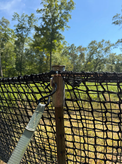 Close-up of a fence with netting and a wooden post, with trees and grass in the background.